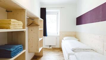 A small, well-lit room with a single bed, wooden shelves, and a window with curtains.
