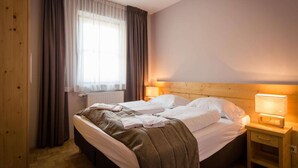 Et hotelværelse med sengegavl i træ, to senge, et natbord med lampe, et vindue med gardiner og en radiator.