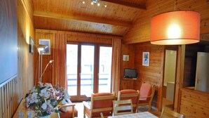 Interior de una cabaña de madera con una mesa de comedor, sillas, un televisor y un ventanal grande.