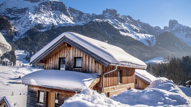 Eine Holzhütte mit geneigtem Dach, umgeben von schneebedeckten Bergen und wolkenlosem Himmel.
