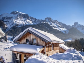A wooden cabin with a sloped roof, surrounded by snow-covered mountains and a clear sky.