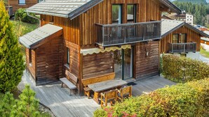 Eine Holzhütte mit Balkon und einer Terrasse mit Sitzgelegenheiten, vor einer Bergkulisse.
