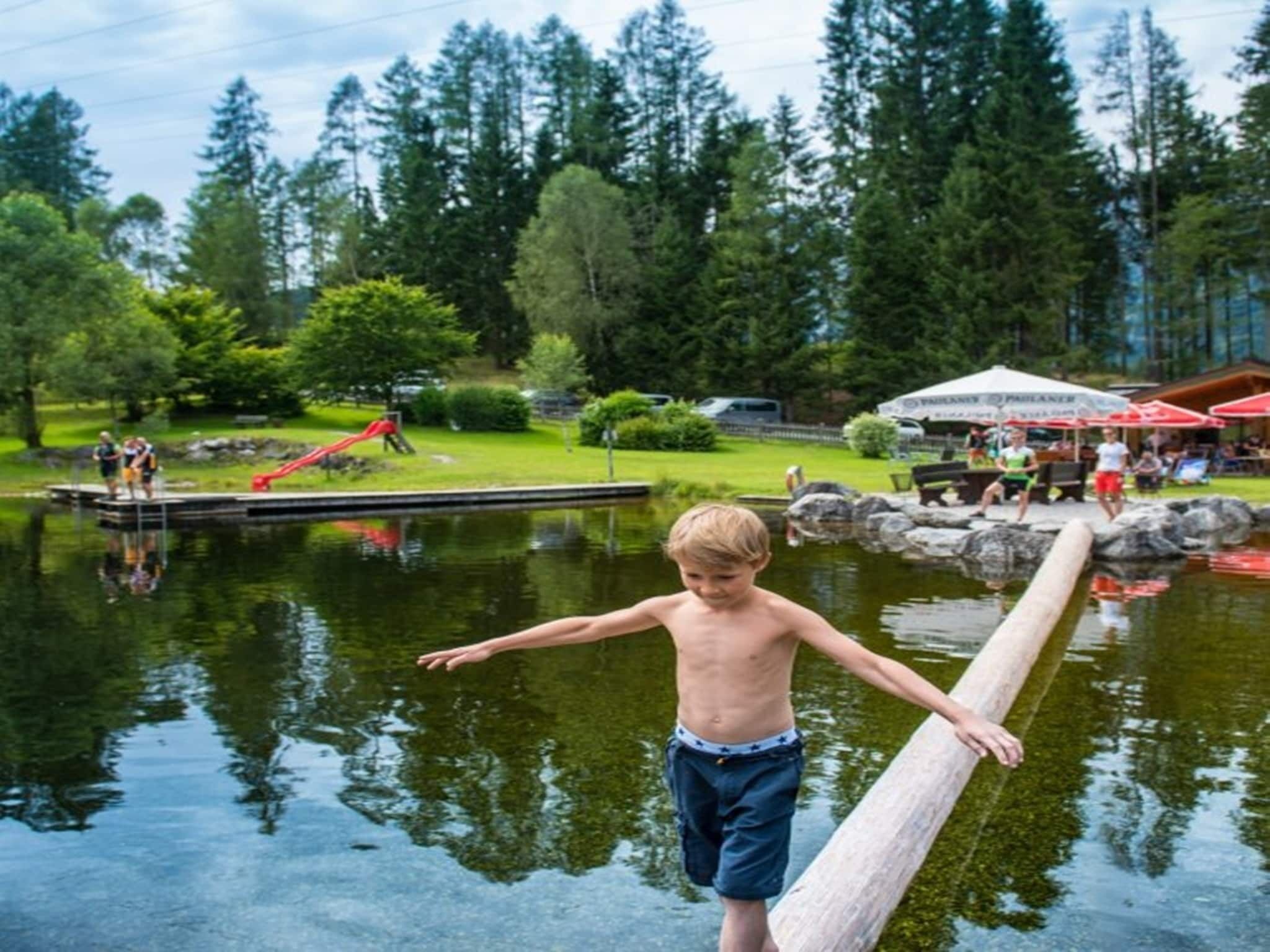 A child balancing on a wooden beam over a body of water, with a playground and trees in the background.