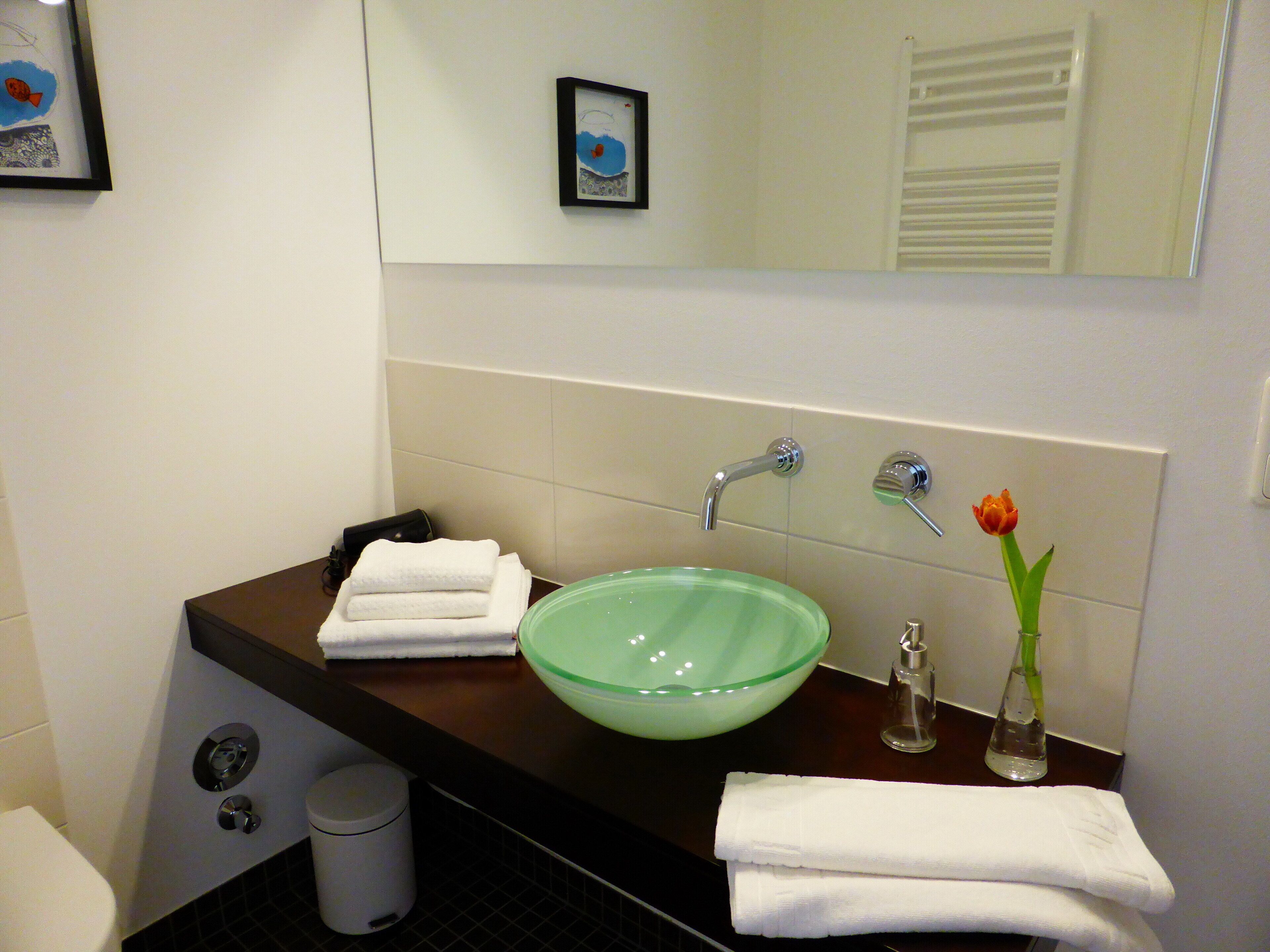 A bathroom with a glass bowl sink, a mirror, a towel rack, and a trash can.