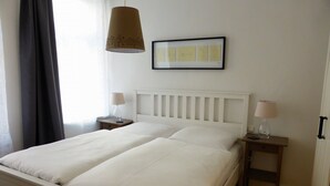 En dobbeltseng med hvide sengetøj, et sengebord med en lampe, og et indrammet billede på væggen.