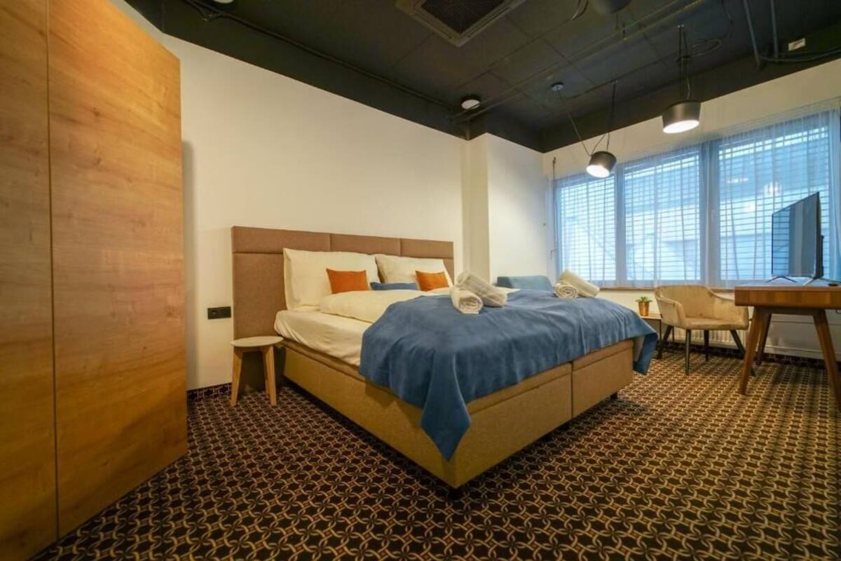 A hotel room with a large bed, a desk, a chair, and a window with blinds.