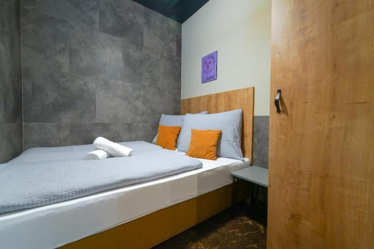 A single bed with a grey and white bedspread, two orange pillows, and a wooden headboard.