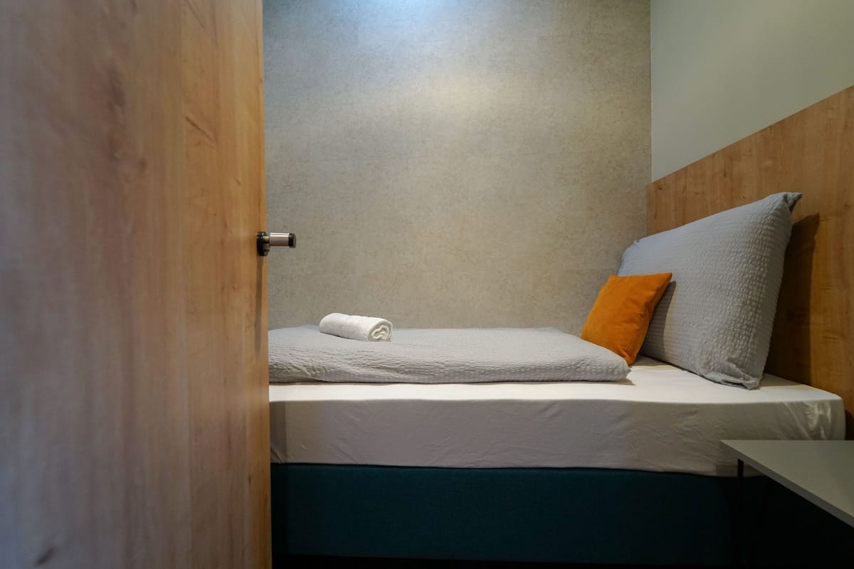 A single bed with a white mattress, grey and orange pillows, and a rolled white towel.