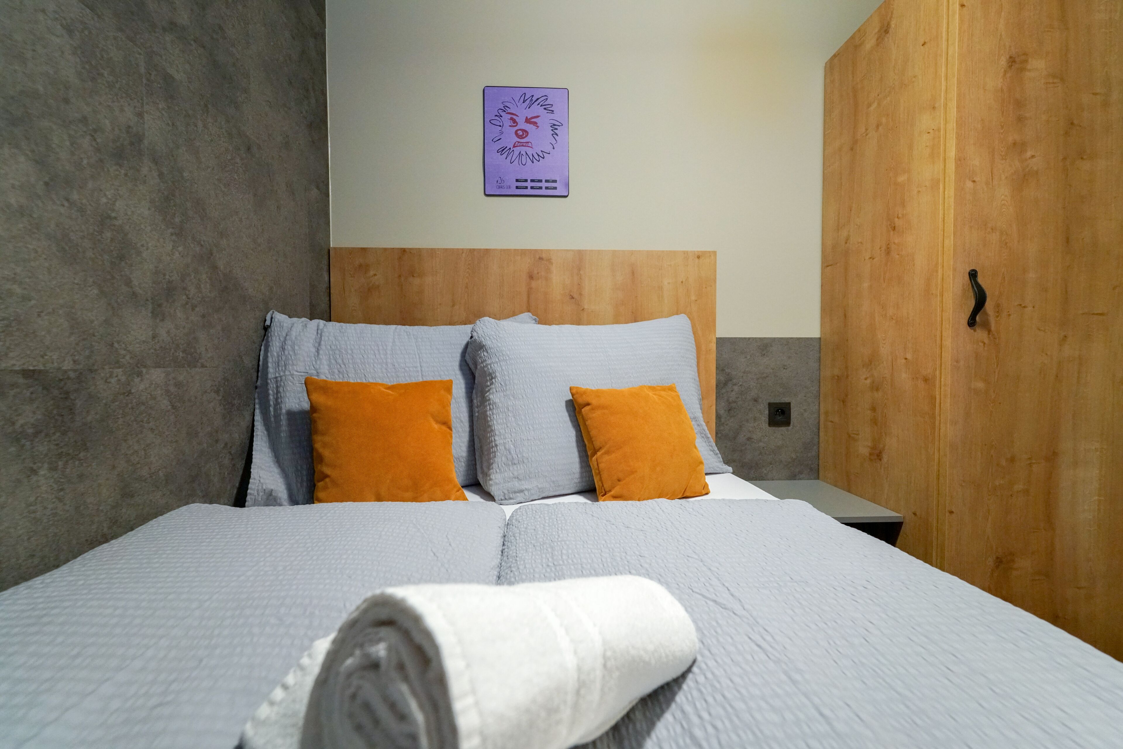 A neatly made bed with a white and gray quilt, two orange pillows, and a white towel on the side.