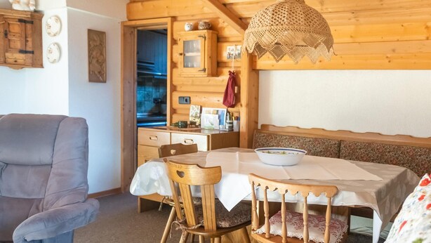 Un interior de madera acogedor con una mesa de comedor, sillas y un banco. Se observa una zona de cocina con armarios y una lámpara colgante.