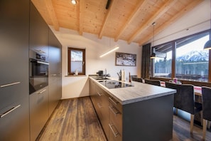 Une cuisine moderne avec des poutres en bois au plafond, un grand îlot central doté d’un évier et une vue sur des montagnes enneigées grâce à de grandes fenêtres.