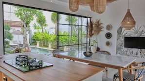 A modern kitchen with a large island, a dining area with a glass table, and a view of a pool and greenery outside. - Good Vibe Villa - 3 Bdrm Luxury Villa (Canggu)