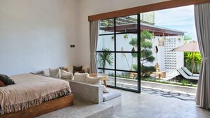 A modern bedroom with a large bed, a sofa, and a view of a garden through a sliding glass door. - Good Vibe Villa - 3 Bdrm Luxury Villa (Canggu)
