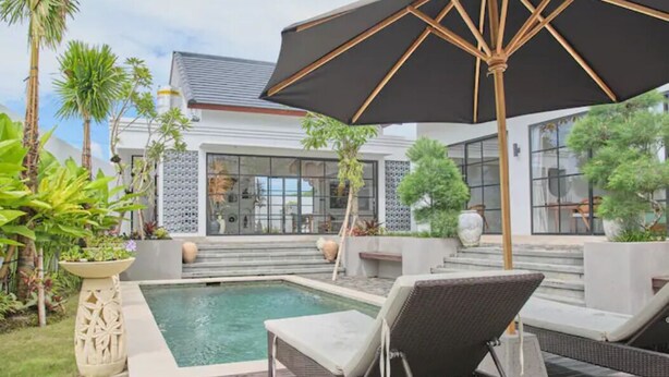 A modern outdoor pool area with lounge chairs and a large umbrella, surrounded by tropical plants and a building with large windows. - Good Vibe Villa - 3 Bdrm Luxury Villa (Canggu)