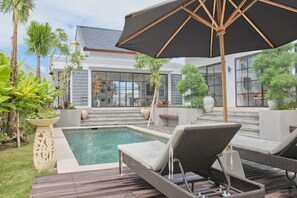 A modern outdoor pool area with lounge chairs and a large umbrella, surrounded by tropical plants and a building with large windows. - Good Vibe Villa - 3 Bdrm Luxury Villa (Canggu)