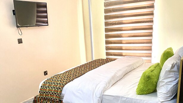 A bedroom with a bed, a TV mounted on the wall, an air conditioning unit, and a window with blinds. - Nue Crest Hotel & Suites (Enugu)