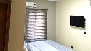 A bedroom with a bed, a window with curtains, an air conditioning unit, and a mounted television. - Nue Crest Hotel & Suites (Enugu)