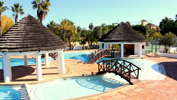 A swimming pool area with two thatched-roof gazebos, a bridge, and palm trees in the background. - Presley Beije Apartment, Quinta do Lago, Algarve (Algarve)