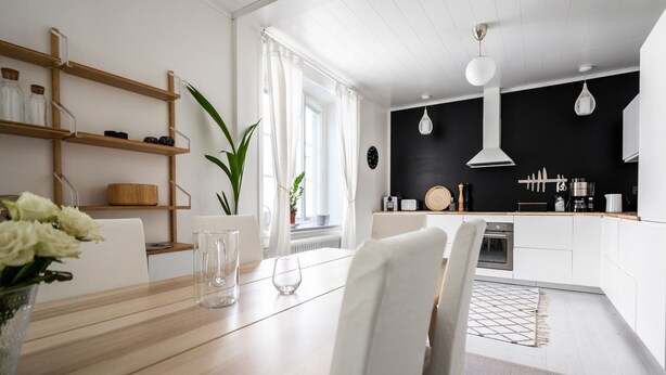 A modern kitchen with white cabinets, a black backsplash, and a wooden countertop. There is a dining area with a wooden table and chairs, and a potted plant on the windowsill. - Luminous 1br next to Puu-Vallilla (Helsinki)
