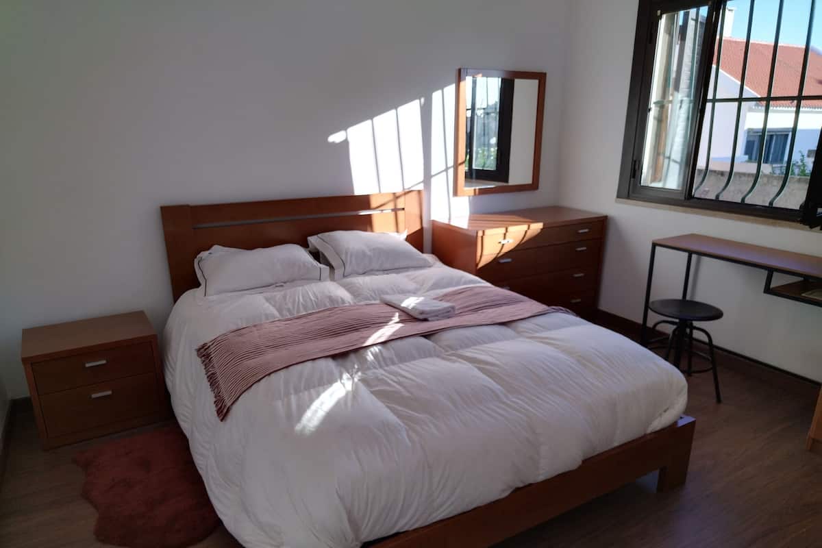 Ein Schlafzimmer mit einem hölzernen Bett, einem Nachttisch, einem Schreibtisch mit Stuhl, einem Spiegel und einem Fenster.