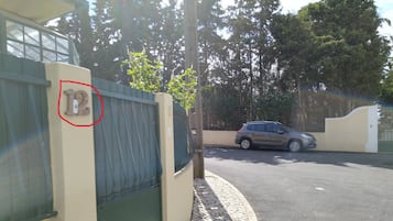 Une entrée fermée par un portail, avec une voiture garée à l’intérieur, un chemin pavé et un bâtiment portant un numéro.