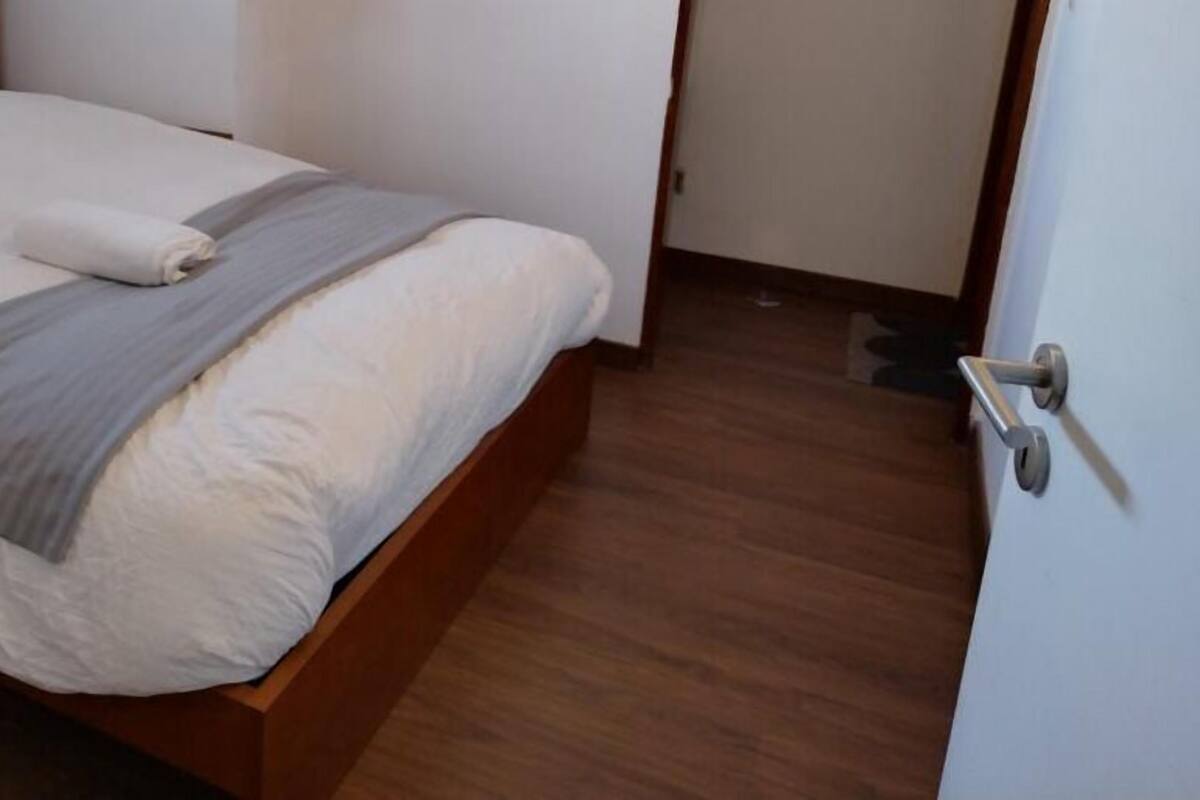 Ein ordentlich bezogenes Bett mit grauer Decke und weißem Kopfkissen, ein hölzernes Kopfteil und eine weiße Tür mit silbernem Griff.