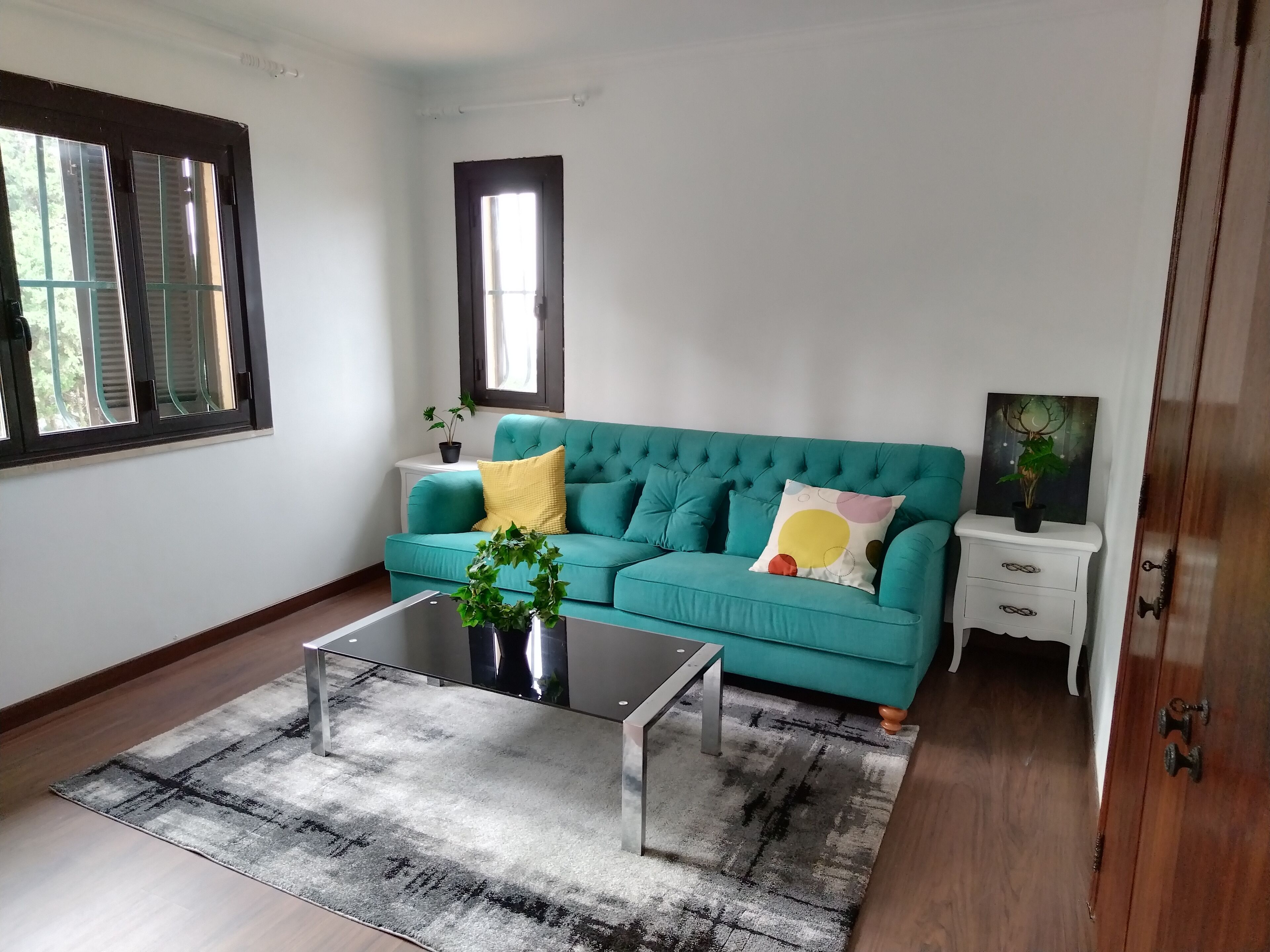 Un sofá color turquesa con cojines decorativos, una mesa de centro de vidrio, una alfombra estampada, una mesita de noche blanca y una puerta de madera.