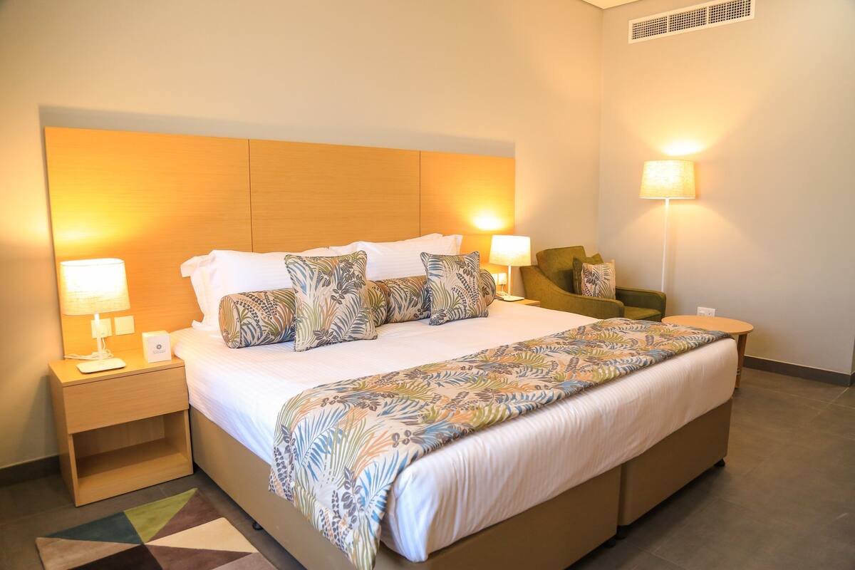 Habitación de hotel con una cama grande, dos mesitas de noche con lámparas, un sillón verde y una manta estampada.