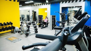 A gym with exercise equipment, a woman using a machine, and a wall with a yellow and blue color scheme. - Ayla Ibri Hotel and Mall (Ibri)