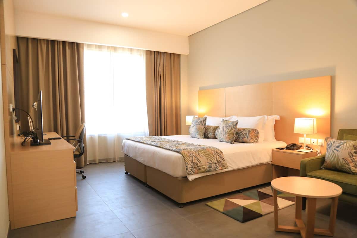 Habitación de hotel con una cama grande, un escritorio, una silla, una mesita pequeña y una mesita de noche con lámpara.
