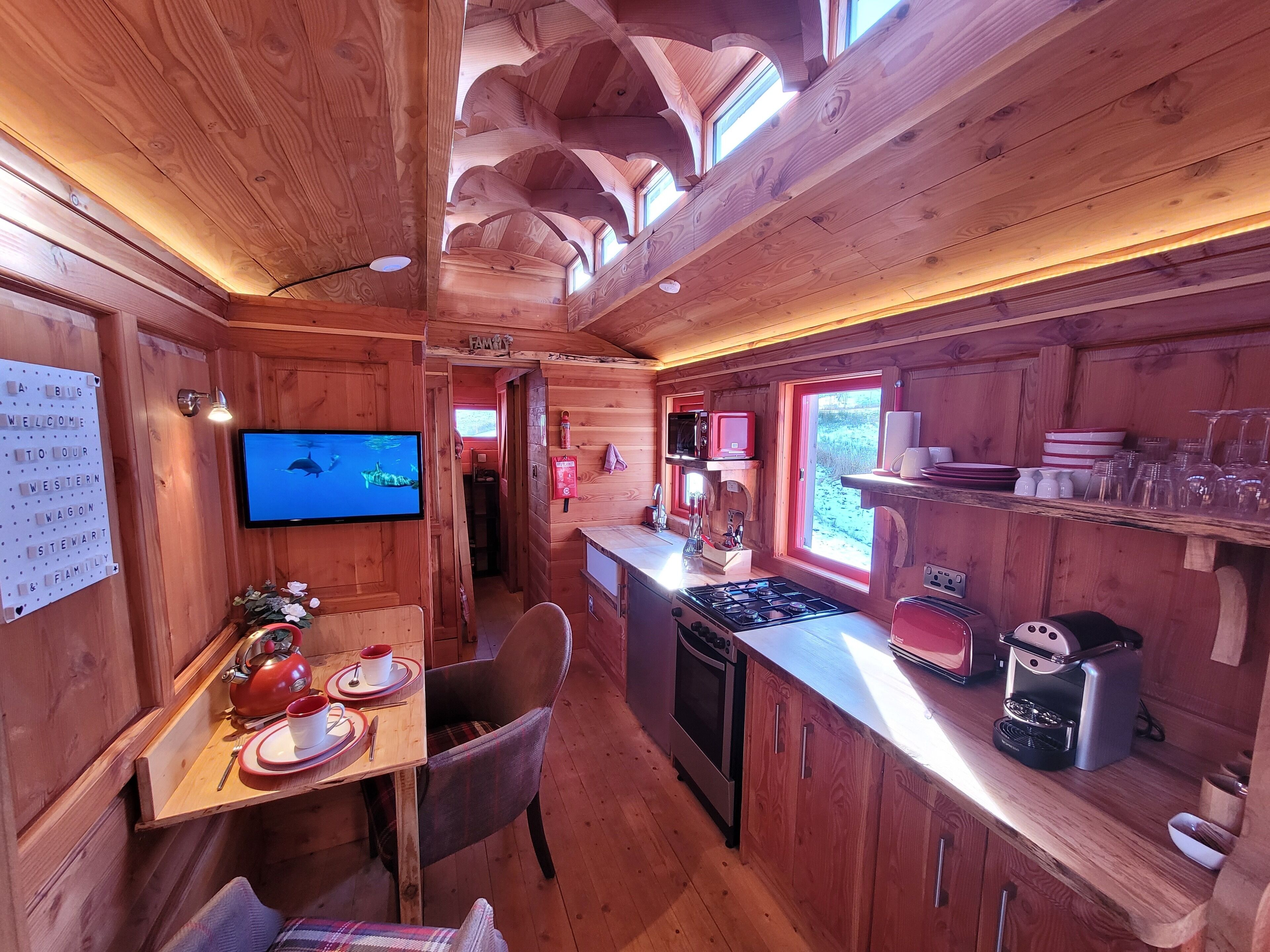 Interior de cabana com decoração em madeira, área de jantar com mesa e cadeiras, cozinha equipada e televisão fixada na parede.