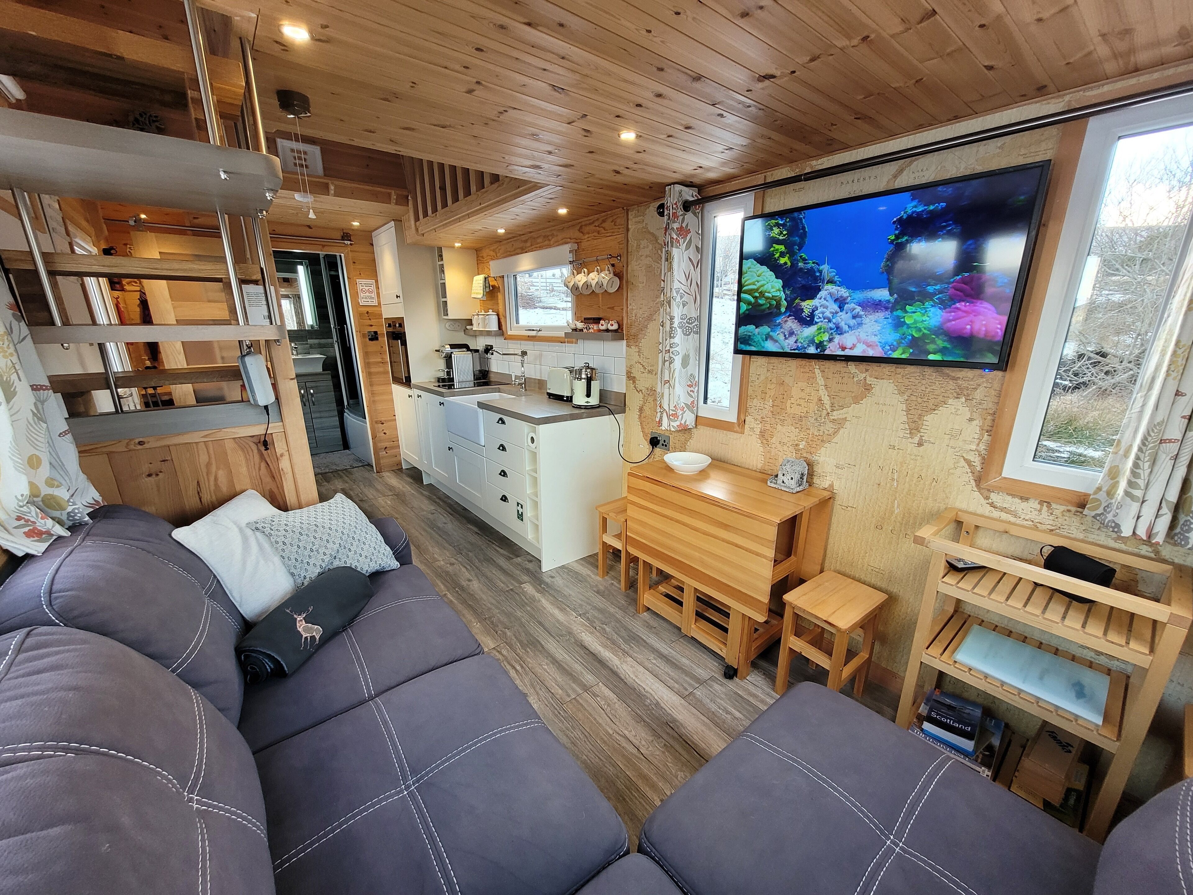 A cozy cabin interior with a sofa, wooden furniture, a kitchen area, and a TV mounted on the wall.