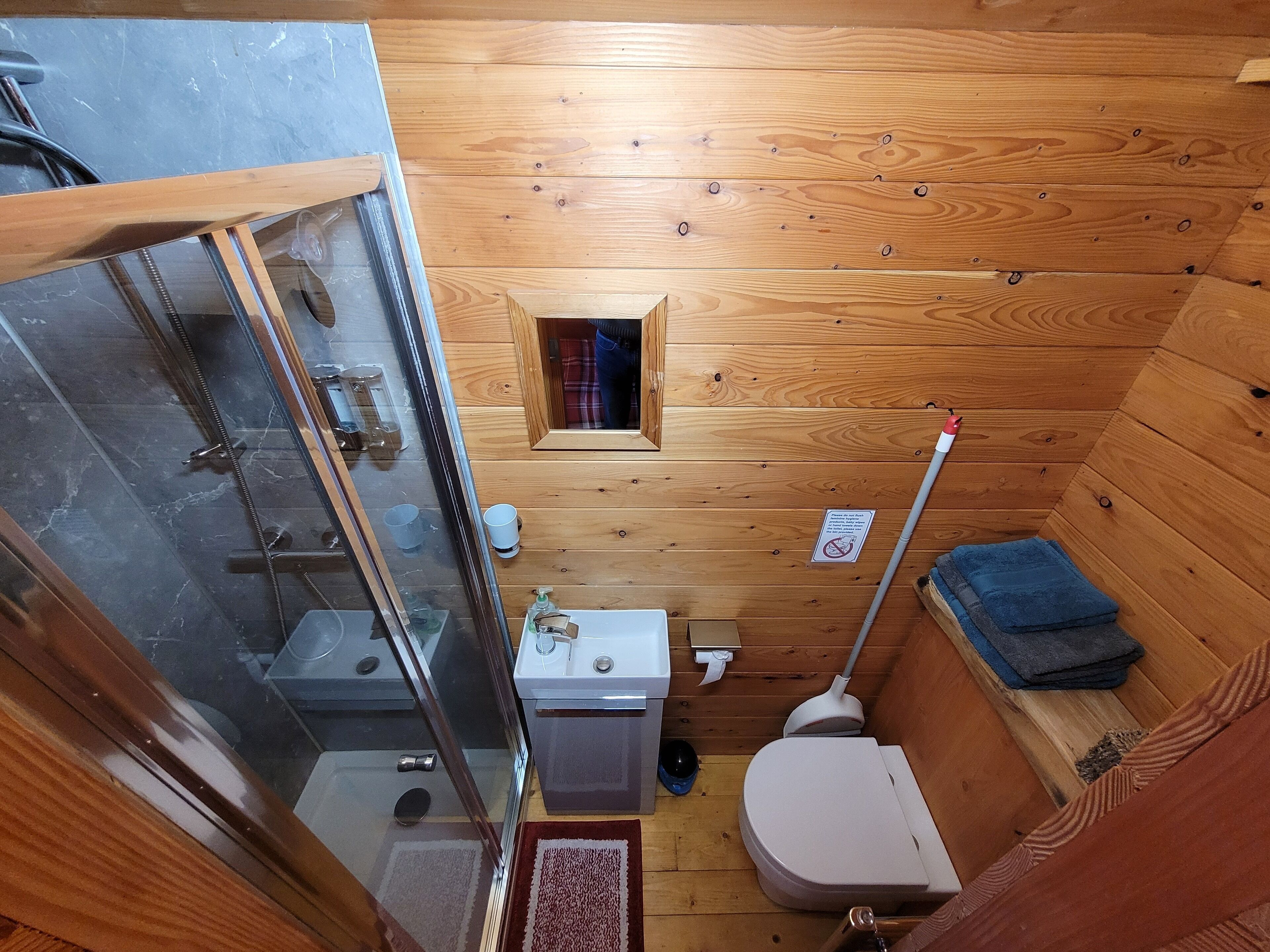 Ein kleines, hölzernes Badezimmer in einer Hütte mit einer Duschkabine aus Glas, einer Toilette, einem Waschbecken und einem kleinen Fenster.