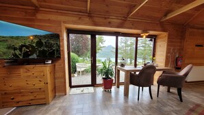 A cozy cabin interior with a wooden ceiling, a dining area with a table and chairs, a television on the left, and a large window offering a view of greenery and a lake.