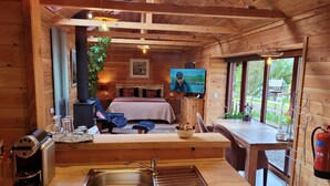 A cozy cabin interior with a wooden interior, a bed with plaid bedding, a television, a dining area, and a kitchen with a coffee maker.