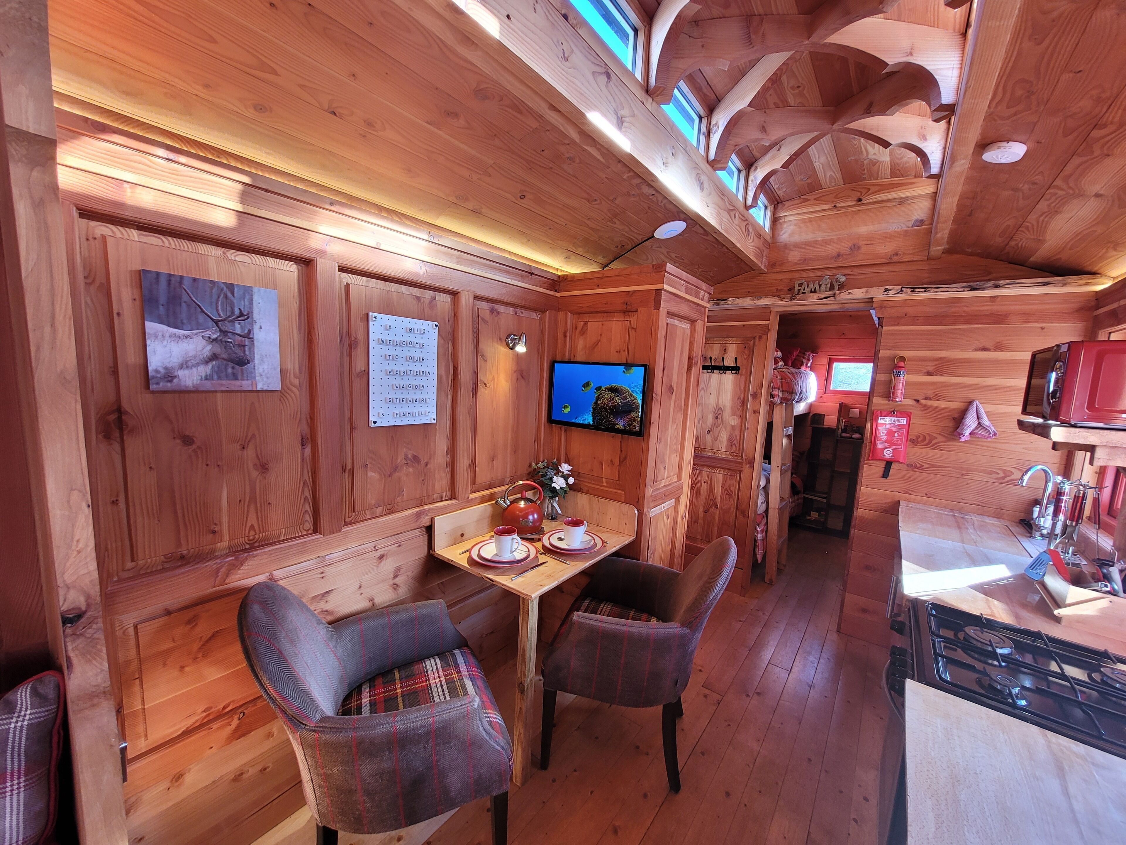 Un intérieur en bois comprenant un coin repas, des chaises, une table avec une théière, un téléviseur et un coin cuisine avec une cuisinière.