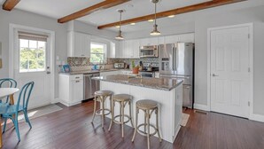 Una cocina con armarios blancos, encimera de granito y electrodomésticos de acero inoxidable. Hay tres taburetes en la isla y vigas de madera en el techo.