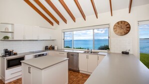 A modern kitchen with white cabinetry, a large island, a view of the sea, and a wooden ceiling. - Surfside - Waterfront with Stunning Views (Culburra Beach)