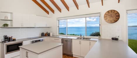 Une cuisine moderne avec des meubles blancs, un grand îlot central, une vue sur la mer et un plafond en bois.