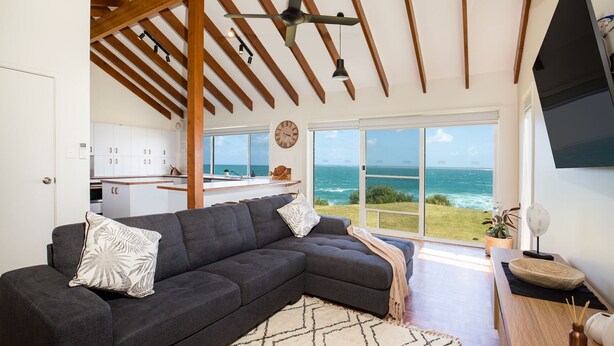 A modern living room with a large gray sectional sofa, a wooden ceiling, and a view of the ocean through large glass doors. - Surfside - Waterfront with Stunning Views (Culburra Beach)