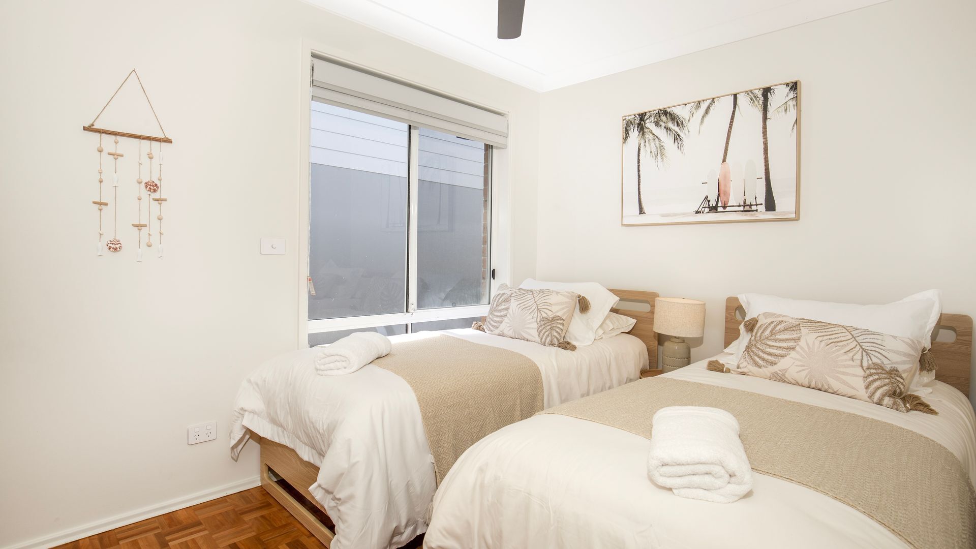 A bedroom with two beds, a window with blinds, a wall-mounted artwork, and a wall-mounted wind chime.