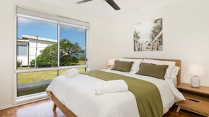 A bedroom with a large window, a bed with white bedding and green pillows, a wooden headboard, a nightstand with a lamp, and a painting of a beach scene. - Surfside - Waterfront with Stunning Views (Culburra Beach)
