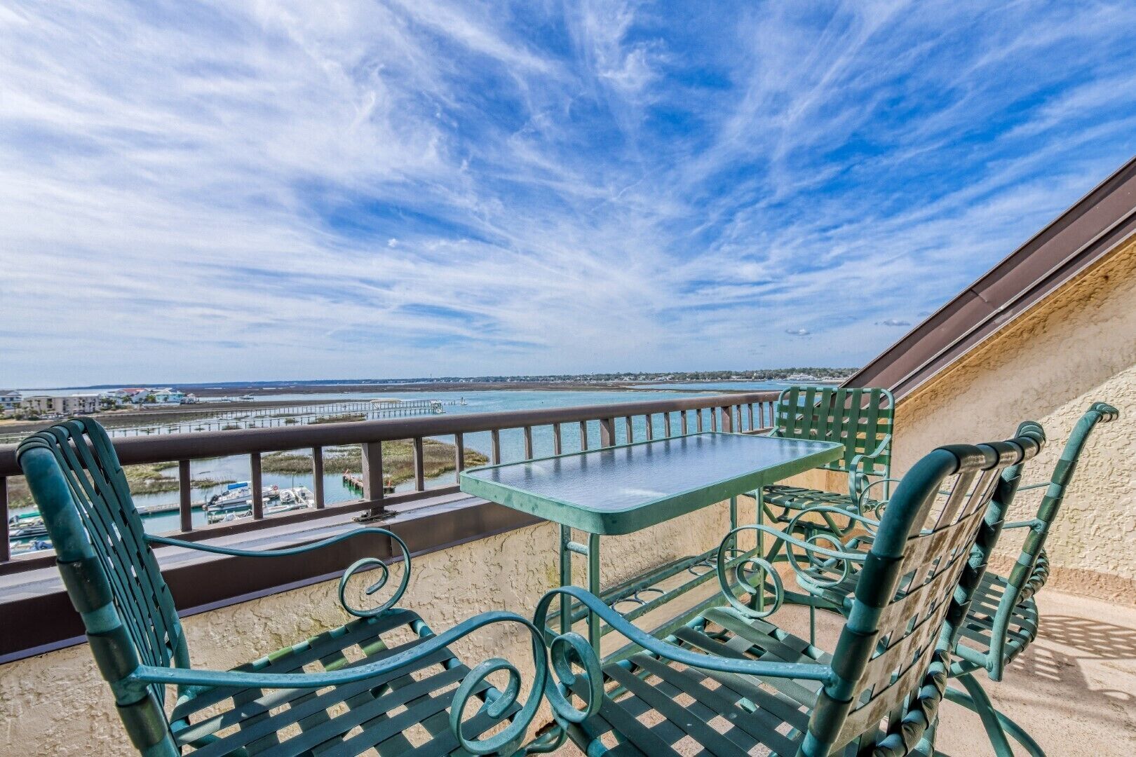 Un balcon doté de mobilier en métal vert, d’une table en verre et offrant une vue sur une zone côtière avec un pont et de l’eau.