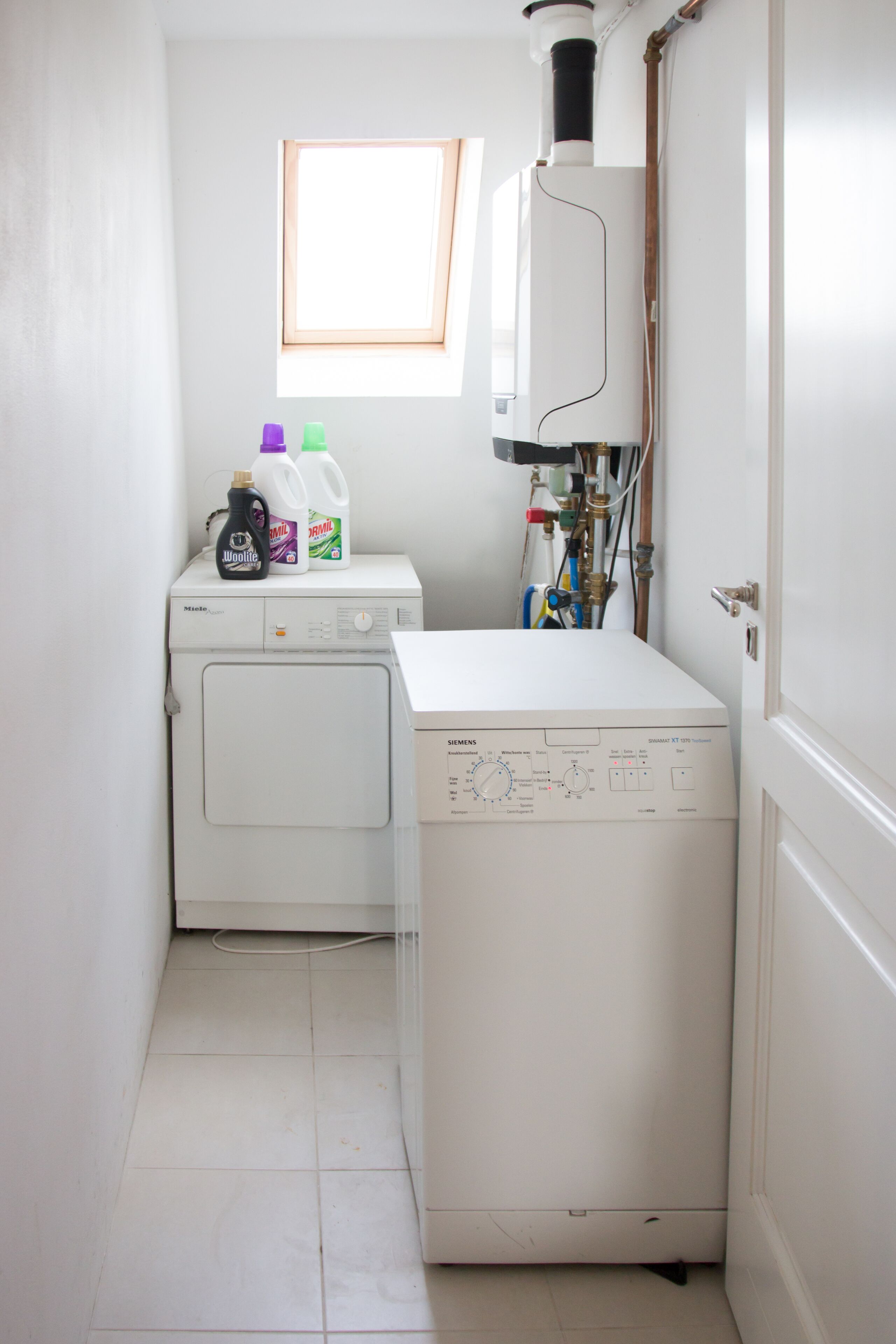 Een wasserette met een wasmachine en droger, een raam en verschillende schoonmaakmiddelen op het aanrecht.