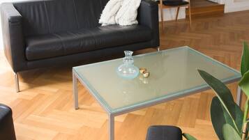 Una sala de estar con un sofá de cuero negro, una mesa de centro de vidrio, una planta en maceta y piso de madera.