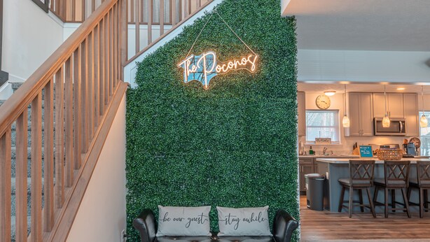 A staircase with wooden railings, a neon sign reading "The Pocanos", and a bench with cushions saying "be our guest" and "stay awhile". - Pocono Getaway with Hot Tub, Games & Fire-pit (Tobyhanna)