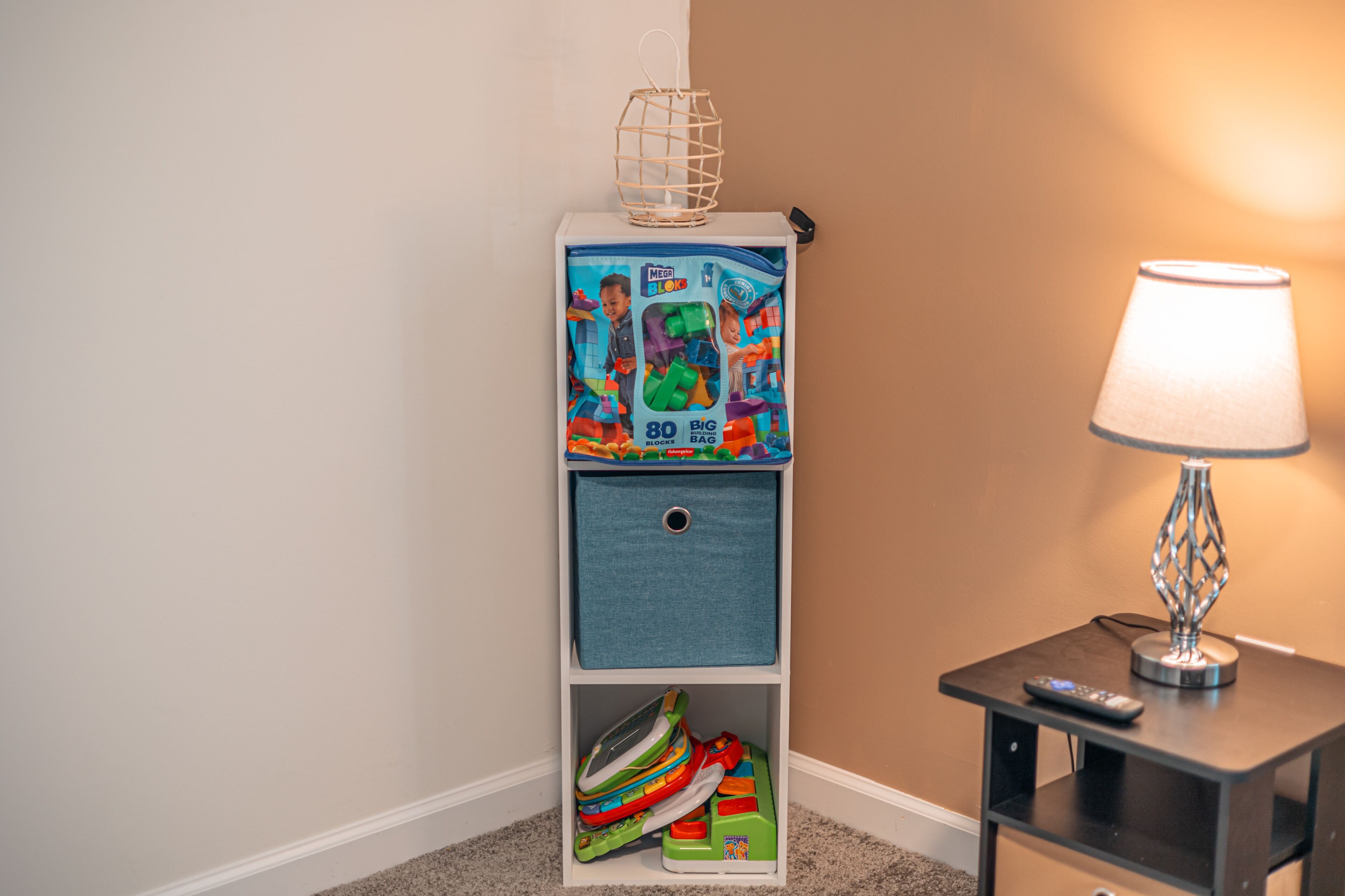 A corner of a room with a white shelf unit containing a Megabolks building block set, a lamp, and a remote control on a side table.