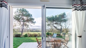 Une pièce dotée d’une porte-fenêtre coulissante donnant sur une terrasse avec une table et des chaises, le tout entouré de verdure et d’arbres.