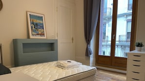 A bedroom with a bed, a bedside table, a dresser, a window with curtains, and a poster on the wall.