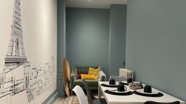 A modern room with a dining table, chairs, a green sofa with yellow and red cushions, and a wall mural of a building.